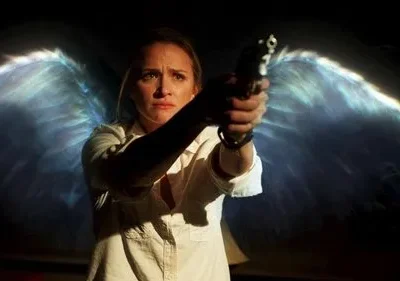 Uma mulher tensa empunha uma arma em um cenário escuro. Duas grandes asas etéreas e brilhantes, com tons de azul e branco, surgem de suas costas.