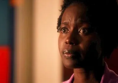 Mulher negra de cabelo crespo, olhos arregalados e boca entreaberta, com expressão de medo ou surpresa. Fundo desfocado, iluminação dramática.