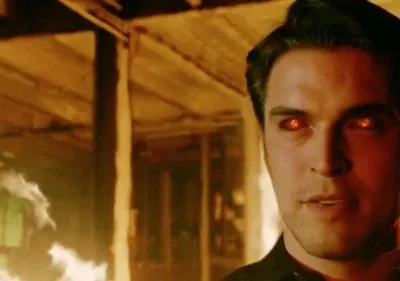 Homem de olhos vermelhos flamejantes encara. Chamas e fumaça laranja iluminam estrutura de madeira em chamas. Clima sombrio e sobrenatural.
