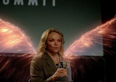 Mulher loira, olhos vermelhos, com asas de energia flamejante. Sentada num palco, com SUMMIT na tela ao fundo. Ambiente sobrenatural.