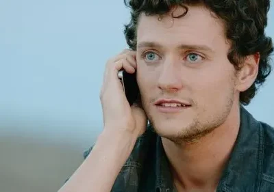 Jovem de cabelo encaracolado e olhos azuis fala ao telefone, com expressão pensativa. Fundo azul claro e desfocado.