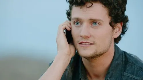 Jovem de cabelo encaracolado e olhos azuis fala ao telefone,com expressão pensativa. Fundo azul claro e desfocado.