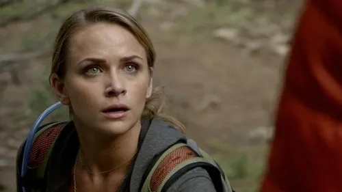 Os Mensageiros – T01E06:Metamorfose 1 Uma mulher loira com mochila e olhar preocupado ou surpreso em um cenário natural. Parte de uma pessoa de laranja à direita.