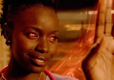 Mulher de pele escura sorri com olhos vermelhos intensos e mão levantada. Fundo laranja embaçado com energia. Clima sobrenatural.