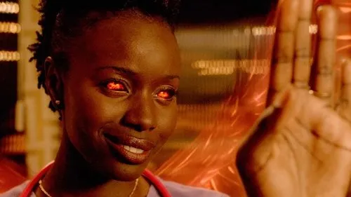 Mulher de pele escura sorri com olhos vermelhos intensos e mão levantada. Fundo laranja embaçado com energia. Clima sobrenatural.