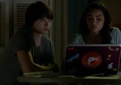 Um jovem e uma jovem olham fixamente para um laptop com adesivos, numa sala escura. O clima é sério e focado.