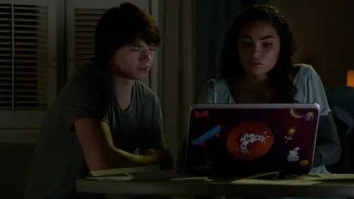 Um jovem e uma jovem olham fixamente para um laptop com adesivos,numa sala escura. O clima é sério e focado.