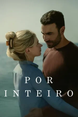 Um casal se abraça suavemente, seus rostos próximos, com o mar azul ao fundo. Ela loira de perfil, ele moreno, com olhar terno. Clima romântico.
