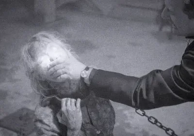 Imagem P&B. Homem com mão na boca de mulher com rosto etéreo e olhos brilhantes. Corrente à vista. Clima sombrio e tenso.