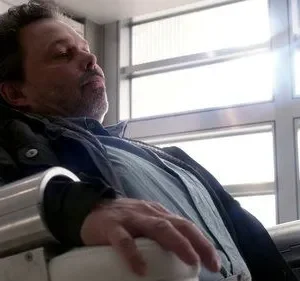Homem de meia-idade com algemas metálicas nos pulsos, sentado em uma cadeira moderna e branca. Ambiente claro e clínico. Parece inconsciente.