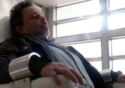 Homem de meia-idade com algemas metálicas nos pulsos, sentado em uma cadeira moderna e branca. Ambiente claro e clínico. Parece inconsciente.