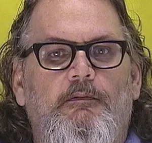 Close-up de um homem de meia idade com óculos e cabelo comprido e grisalho, contra um fundo amarelo. Ele parece sério e um pouco sombrio.