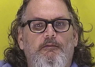 Close-up de um homem de meia idade com óculos e cabelo comprido e grisalho, contra um fundo amarelo. Ele parece sério e um pouco sombrio.