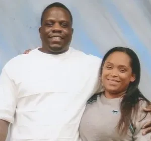 Claro, aqui está um texto alternativo conciso para a imagem: Um casal de pele escura está abraçado em um estúdio de fotos. O homem usa uma camisa branca, enquanto a mulher está usando uma camiseta bege. Um fundo de estúdio azul pálido está atrás deles. A foto parece formal.