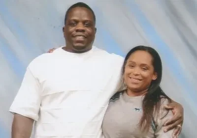 Claro, aqui está um texto alternativo conciso para a imagem: Um casal de pele escura está abraçado em um estúdio de fotos. O homem usa uma camisa branca, enquanto a mulher está usando uma camiseta bege. Um fundo de estúdio azul pálido está atrás deles. A foto parece formal.