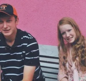Dois adolescentes sentados em um banco contra uma parede rosa. Um menino usa um boné de beisebol e camisa listrada. Uma garota com cabelo ruivo longo está sentada ao lado dele, sorrindo para a câmera. O clima é casual e amigável.