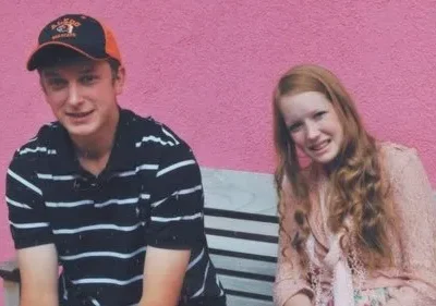 Dois adolescentes sentados em um banco contra uma parede rosa. Um menino usa um boné de beisebol e camisa listrada. Uma garota com cabelo ruivo longo está sentada ao lado dele, sorrindo para a câmera. O clima é casual e amigável.