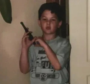 Um jovem rapaz, usando uma camiseta de futebol, segura um walkie-talkie perto de uma porta interior. A cena é íntima e aparentemente doméstica.