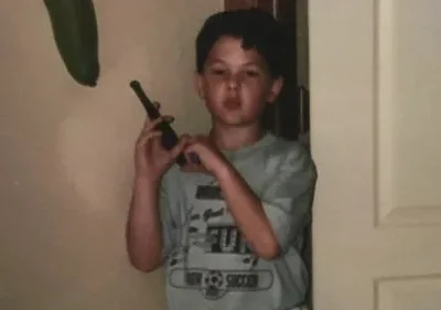 Um jovem rapaz, usando uma camiseta de futebol, segura um walkie-talkie perto de uma porta interior. A cena é íntima e aparentemente doméstica.