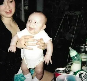 Claro. Aqui está uma descrição concisa da imagem: Um bebê sorridente é mantido por uma mulher enquanto um homem de chapéu olha para ele. Fundo escuro e bagunçado com bebidas e uma mesa. Cena familiar e aconchegante.