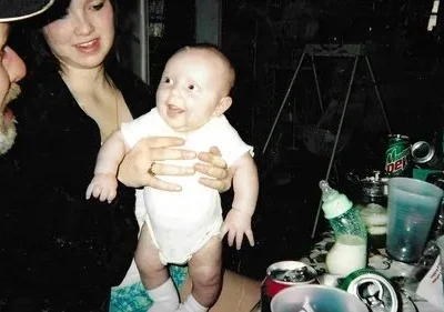 Claro. Aqui está uma descrição concisa da imagem: Um bebê sorridente é mantido por uma mulher enquanto um homem de chapéu olha para ele. Fundo escuro e bagunçado com bebidas e uma mesa. Cena familiar e aconchegante.