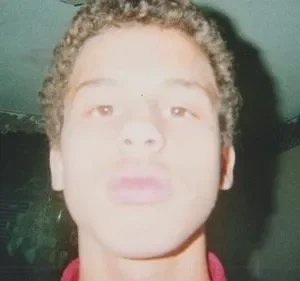 Claro, aqui está uma legenda alternativa para a imagem: Close-up de um jovem, rosto levemente desfocado, em uma sala mal iluminada. Paredes verde-acinzentadas com um rodapé decorado. Clima íntimo e nostálgico.
