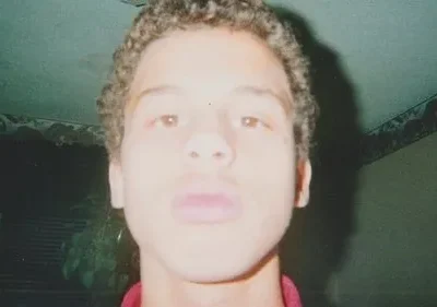 Claro, aqui está uma legenda alternativa para a imagem: Close-up de um jovem, rosto levemente desfocado, em uma sala mal iluminada. Paredes verde-acinzentadas com um rodapé decorado. Clima íntimo e nostálgico.