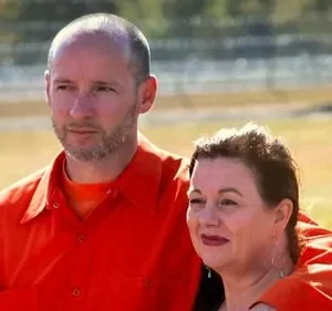 Dois presos em macacões laranja, um homem e uma mulher, abraçados em um pátio de prisão ensolarado. Um olhar pensativo nos seus rostos.