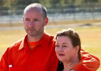 Dois presos em macacões laranja, um homem e uma mulher, abraçados em um pátio de prisão ensolarado. Um olhar pensativo nos seus rostos.
