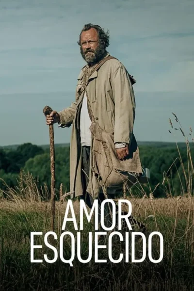 Homem barbudo e desalinhado com casaco bege e cajado, parado em um campo gramado com floresta e céu nublado ao fundo. Clima rural.