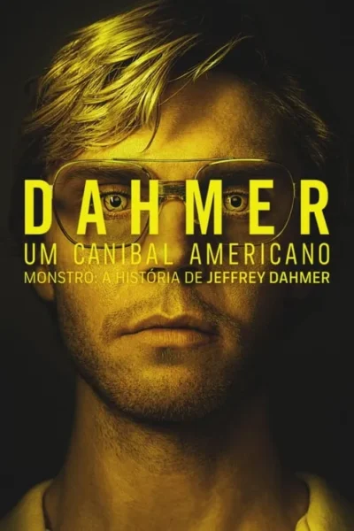 Close-up de um homem com óculos aviador, cabelo loiro bagunçado e expressão séria. Iluminação amarelada contra fundo escuro e sombrio.