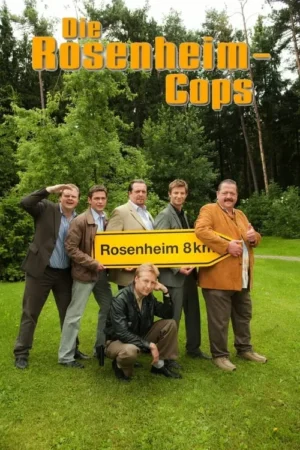 Seis homens em roupas sociais casuais posam em um gramado verde com floresta ao fundo. Quatro seguram uma placa de direção amarela. Clima divertido.