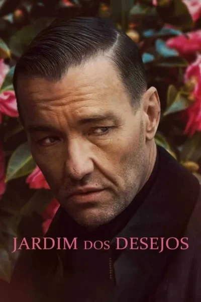 Close-up de um homem sério com cabelo escuro e barba por fazer, olhando para baixo. Fundo borrado de folhas verdes e flores cor-de-rosa.