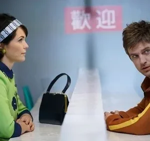 Mulher de vestido mod verde e homem de jaqueta laranja se sentam frente a frente em balcão, letreiro asiático ao fundo. Clima retrô e misterioso.