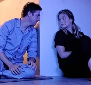 Homem de pijama e mulher em roupa escura se encaram sentados no chão, sob luz azul. Rostos tensos, clima de apreensão.