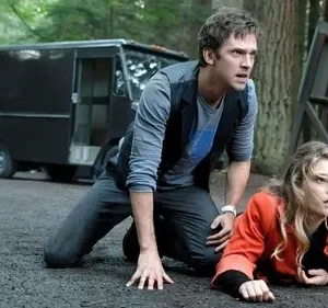 Um homem de joelhos e uma mulher loira deitada, ambos apavorados numa estrada de terra na floresta. Van escura no fundo.