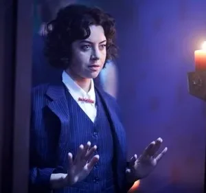 Mulher de terno listrado e cabelo encaracolado olha tensa, mãos erguidas. Velas acesas em ambiente escuro criam clima misterioso.