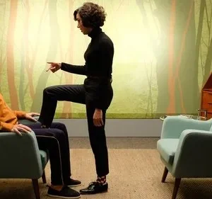 Uma mulher de preto, com pé no colo e dedo em riste, confronta um homem sentado em poltrona azul. Fundo peculiar de floresta estilizada.