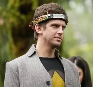 Jovem com aparelho tecnológico na testa, casaco cinza e camiseta preta com triângulo amarelo, olhando pensativo. Fundo verde.