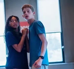 Uma garota e um garoto adolescentes, ele loiro, olham assustados em um ambiente interno. A garota tem cabelos cacheados. Clima tenso.