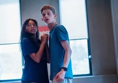 Uma garota e um garoto adolescentes, ele loiro, olham assustados em um ambiente interno. A garota tem cabelos cacheados. Clima tenso.