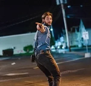 Homem barbudo de camisa azul apontando um rádio, inclinando-se em rua escura à noite. Prédios ao fundo, clima de urgência.