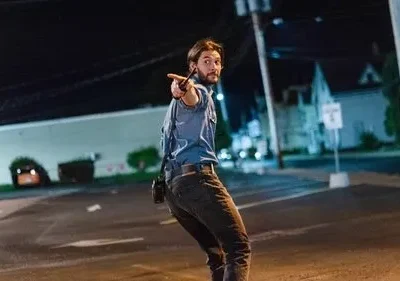 Homem barbudo de camisa azul apontando um rádio, inclinando-se em rua escura à noite. Prédios ao fundo, clima de urgência.