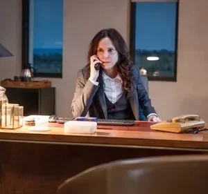 Mulher séria em terno fala ao telefone em uma escrivaninha de escritório iluminada. Janelas mostram a noite; clima profissional e tenso.