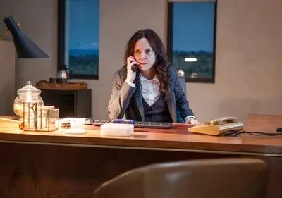 Mulher séria em terno fala ao telefone em uma escrivaninha de escritório iluminada. Janelas mostram a noite; clima profissional e tenso.