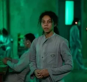 Jovem mulher em uniforme cinza olha intensamente. Pessoas uniformizadas sentadas ao fundo em sala com forte luz verde e clima sombrio.