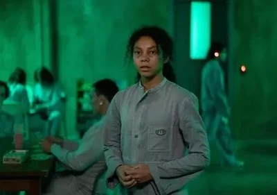 Jovem mulher em uniforme cinza olha intensamente. Pessoas uniformizadas sentadas ao fundo em sala com forte luz verde e clima sombrio.