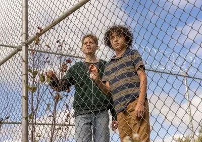 Um homem e um menino olham para cima por uma cerca de arame com arame farpado, contra o céu azul e nublado. Clima de anseio.