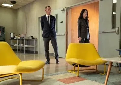 Um homem de terno e uma mulher de blazer sérios em uma sala de espera moderna com cadeiras amarelas, máquina de venda e um mural colorido.