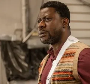 Homem negro de camisa vinho, colete estampado e tipoia, olha com apreensão. Fundo claro, ambiente interno simples.